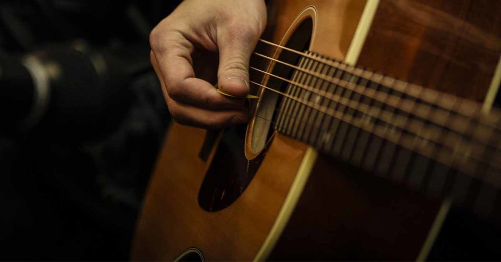 Image of an acoustic brown guitar, a hand can be seen holding a plectrum.