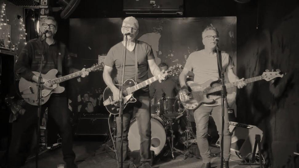 Black and white photo of four band members performing live on stage with bass guitar, two electric guitars and drums, with three members singing.
