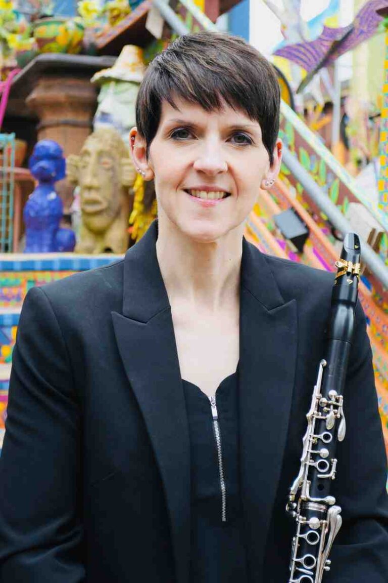 Amanda Morrison wearing a black jacket and top with a clarinet visible and a multicolour artistic background behind her