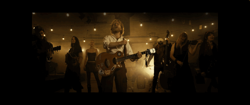 Eight band members, four women and four men, on stage holding guitars, double bass, violin, and accordion, bathed in warm yellow stage lighting, all facing forward.