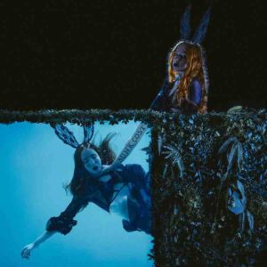 RXRXBBIT featured in two contrasting scenes, one with her peeking through a hedge and the other underwater, wearing rabbit ears. The overall tone is dark and moody, matching the edgy style of the cover art for her track Sunk Cost.