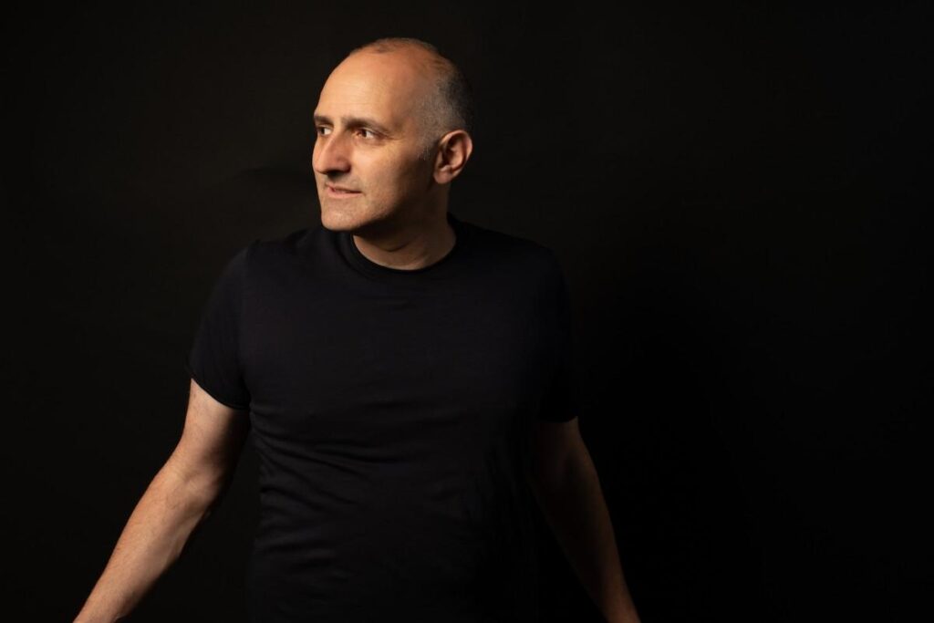 Eyal Erlich wearing a black T shirt against a dark background, looking slightly off to the side.