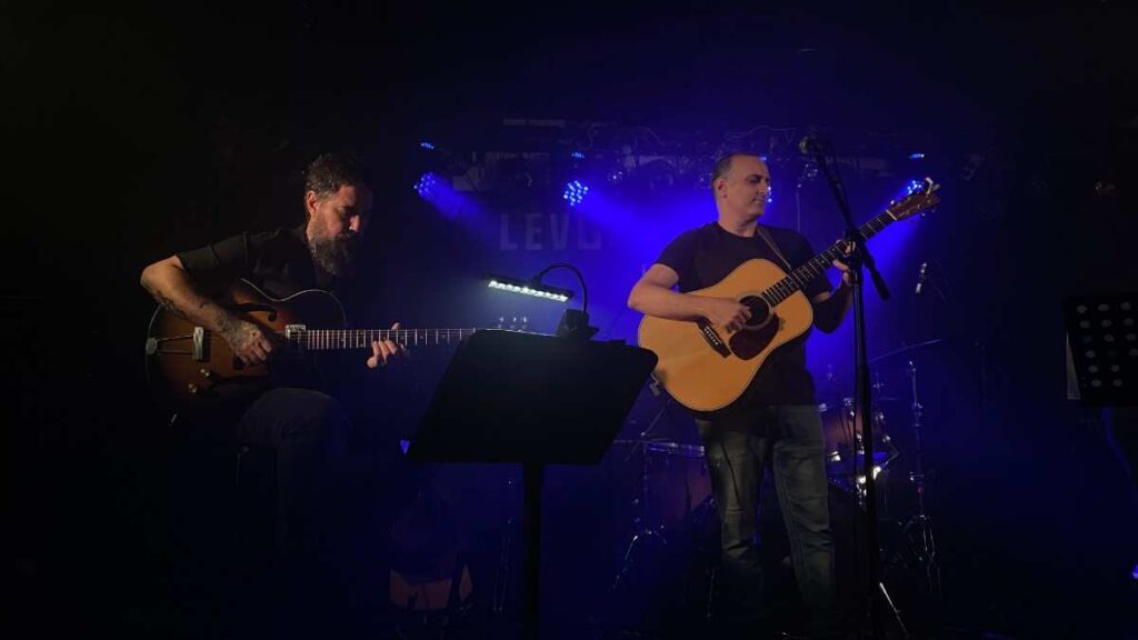 Eyal Erlich performing live on stage under blue lighting with an acoustic guitar, accompanied by an electric guitarist seated beside him and a drum kit visible in the background.