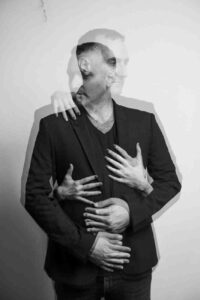Black and white blurred portrait of Eyal Erlich in a black suit and T shirt with his hand on his chest and multiple ghosted images around him.