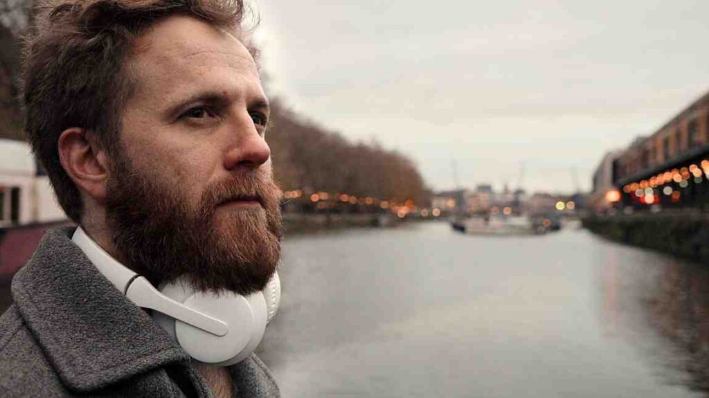 Close up of composer Joe Galuszka wearing white headphones by the river in Bristol, captured from the left side with soft late afternoon light.