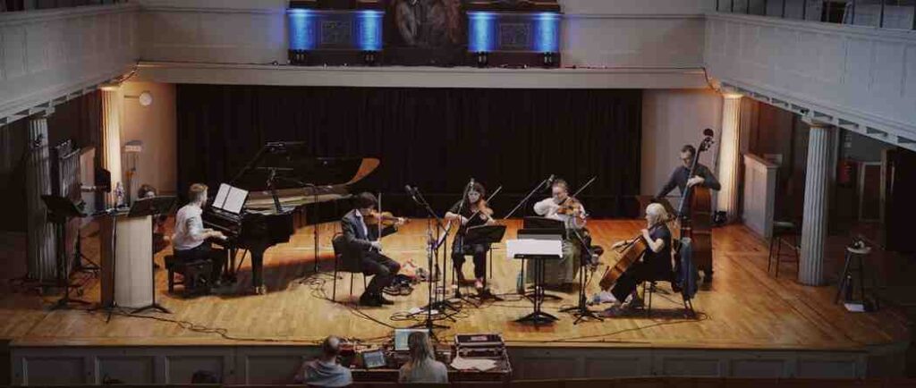Full ensemble performing on the recording stage at St George’s Bristol with Joe Galuszka on piano and string players arranged in a traditional orchestral layout viewed from above in warm soft lighting.