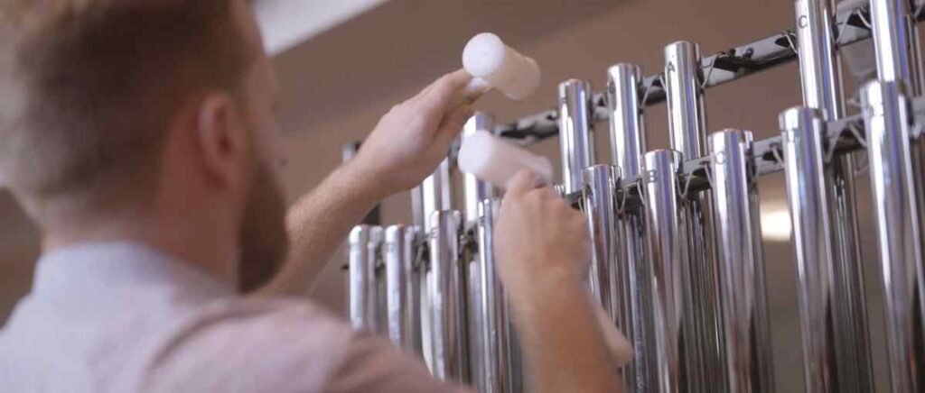 Composer Joe Galuszka playing tubular bells during a recording session, holding two plastic hammer beaters while performing on a set of C major bells with his back turned slightly toward the camera.