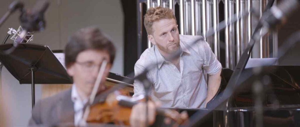 Composer Joe Galuszka standing behind a focused violinist during a recording session with a blurred mic stand in the foreground showing the studio environment.