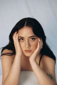 Tia Gostelow lying on a white background, looking confidently at the camera with her hands cupping her face.