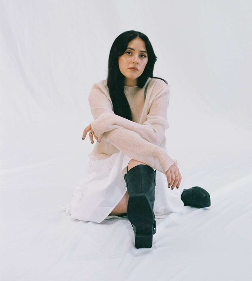Tia Gostelow sitting on the floor with white draped material behind her, wearing a pale salmon cardigan and black boots, legs and arms crossed, looking confidently at the camera.