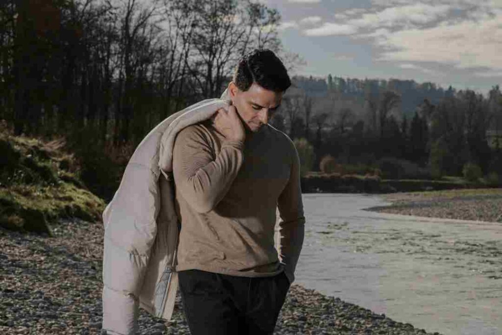 Artist Gabriele Rizzo standing by a river with a pebble shoreline and bare winter trees, looking down in a reflective pose with a puffy jacket over his shoulder.