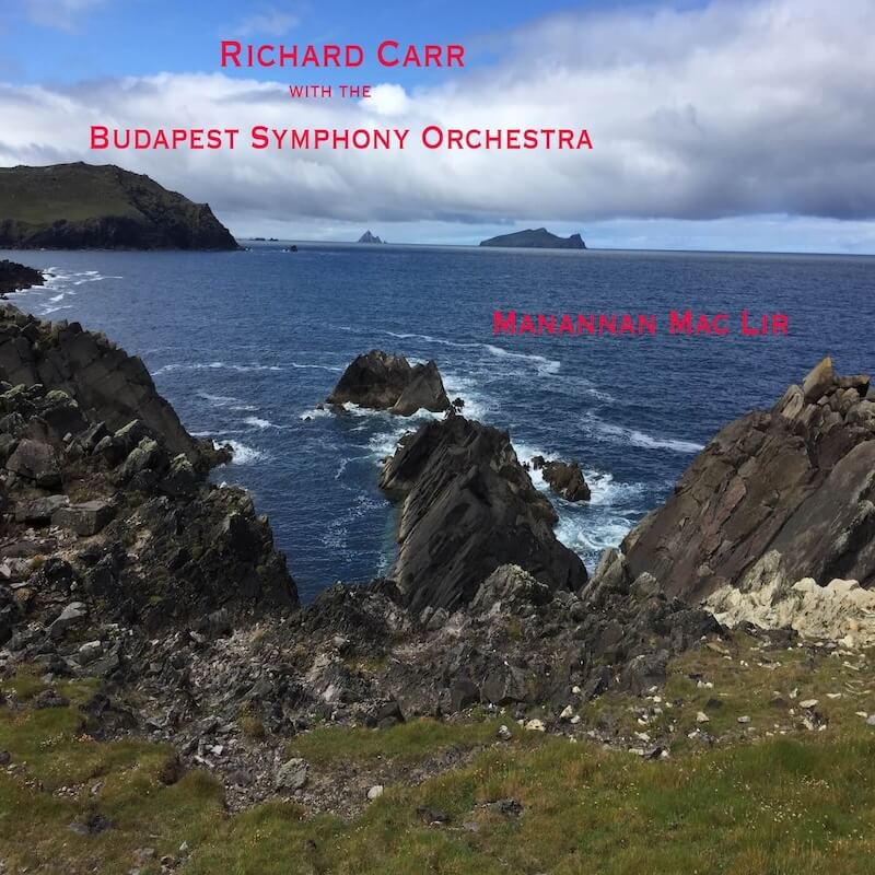 Island coastline with rugged cliffs and rocks, waves breaking against the shore, wild grass in the foreground, and storm clouds over the sea for Richard Carr Manannan mac Lir album cover