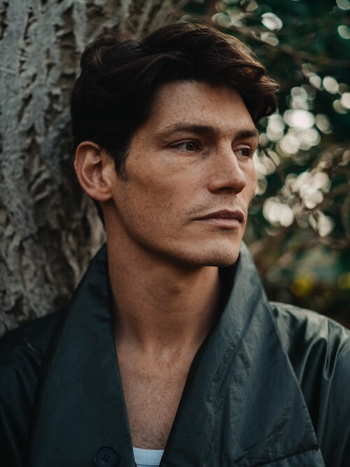 Sam Way resting against a tree outdoors, light falling on his face from the right, contemplative headshot of independent artist.