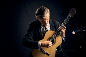 Neno Munitic holding a classical guitar in performance position against a black background, wearing a suit and tie and looking down in focused concentration