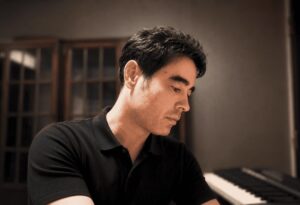 Portrait of composer and pianist Charles Knudsen wearing a black polo shirt with a piano in the background. He looks down thoughtfully to his left against a light grey backdrop, reflecting the contemplative nature of his contemporary classical music.