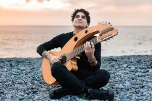Daniele Fabio sitting cross legged on a Mediterranean beach playing harp guitar, dressed in black and looking peaceful as he performs beside the sea.