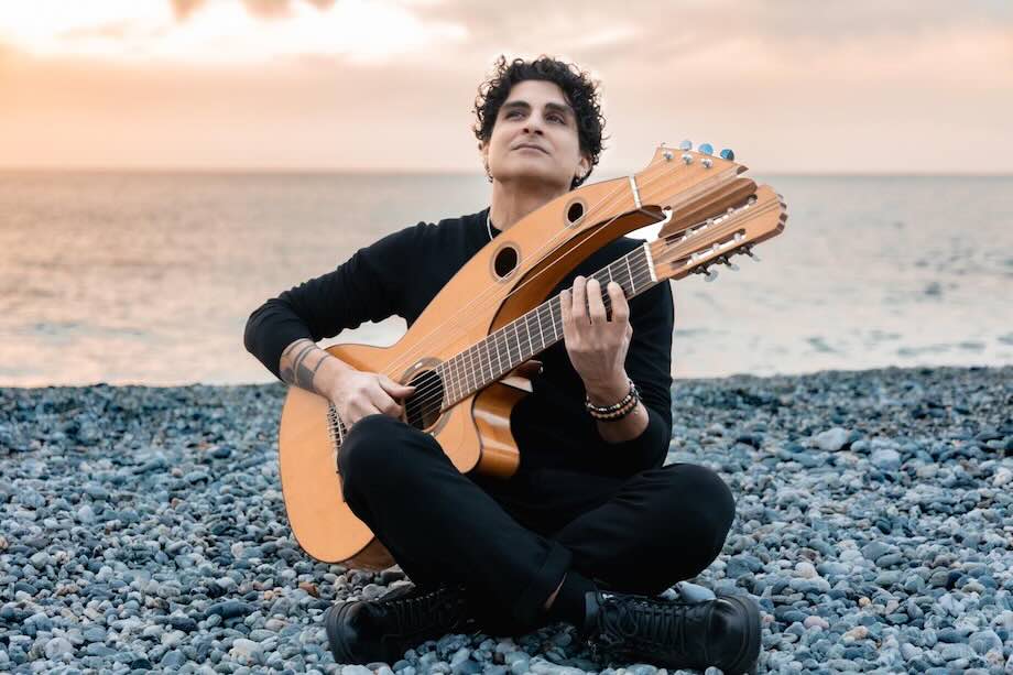 Daniele Fabio sitting cross legged on a Mediterranean beach playing harp guitar, dressed in black and looking peaceful as he performs beside the sea.