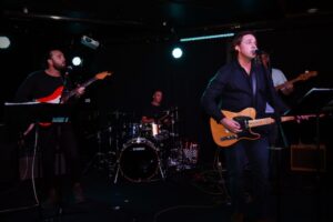 FINN performing Still Believe live on stage with four band members under warm stage lighting, featuring acoustic rock instrumentation and an intimate live performance.