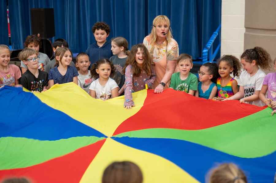 Kim Noller stands at the centre of a group of children holding a large colourful colour wheel fabric while she speaks to them through a microphone.